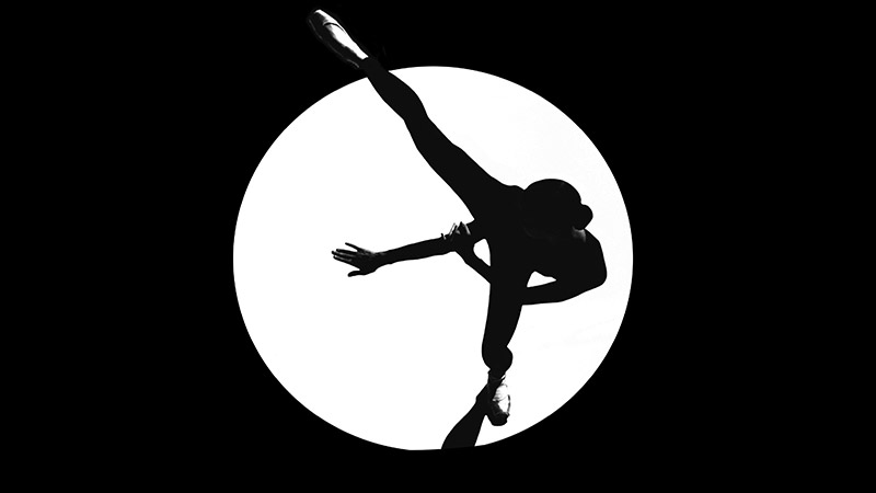 a black and white silhouette of a ballet dancer in a spotlight extending her leg in to the side and her hands forward