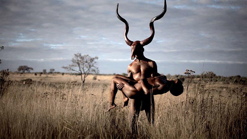 a man wearing a skull from an animal with large, spiraled antlers carries the limp body of another man in a grassy landscape
