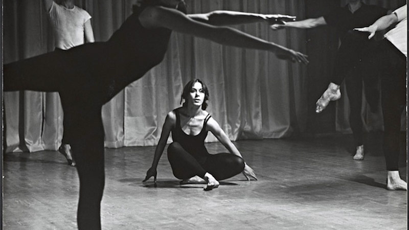 a historic photo of dancer/choreographer Yvonne Rainer from the 1960s in which she is seated with legs crossed, framed by the legs and feet of other dancers, all in black dancewear