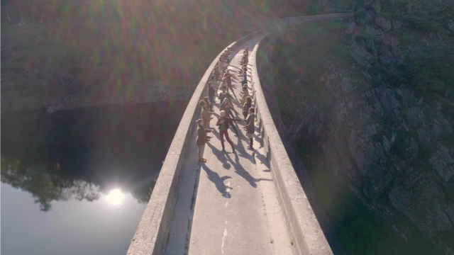 an aerial shot of two dozen people dancing in unison on top of a concrete dam