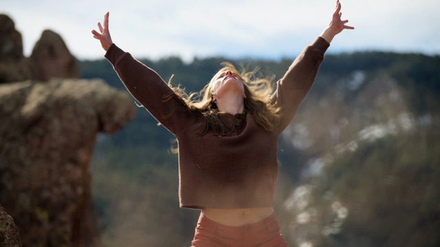 above a shallow valley with moss rocks around, a light skinned dancer in a brown sweatshirt opens her chest and arms to the sunlight