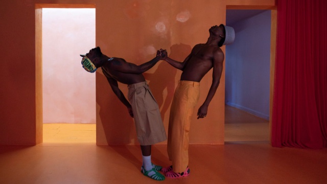 in a empty, clean house, two shirtless dark skinned men clasp their right hands, using each other's strength to support themselves as they each lean backward